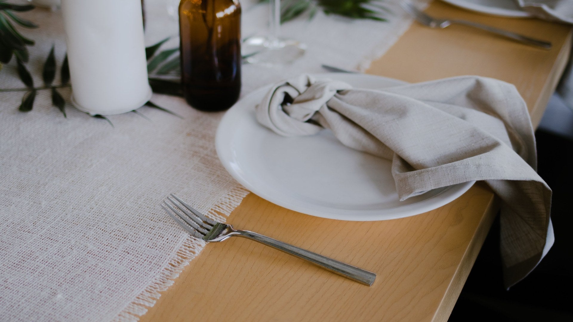 A woven cotton table runner laid down a wooden table with a linen napkin, plate and fork beside its fringed edge.