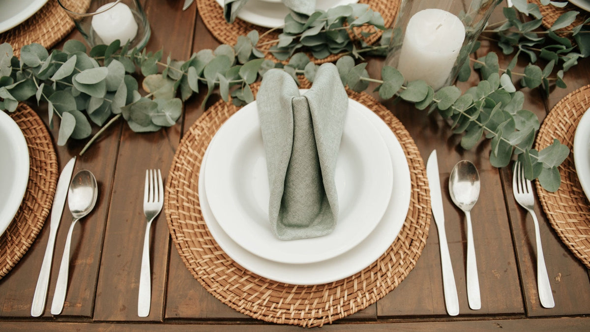 A place setting seen from above - a sage green cloth napkin folded on stacked white plates over a round woven placemat, with cutlery on a dark wood table.