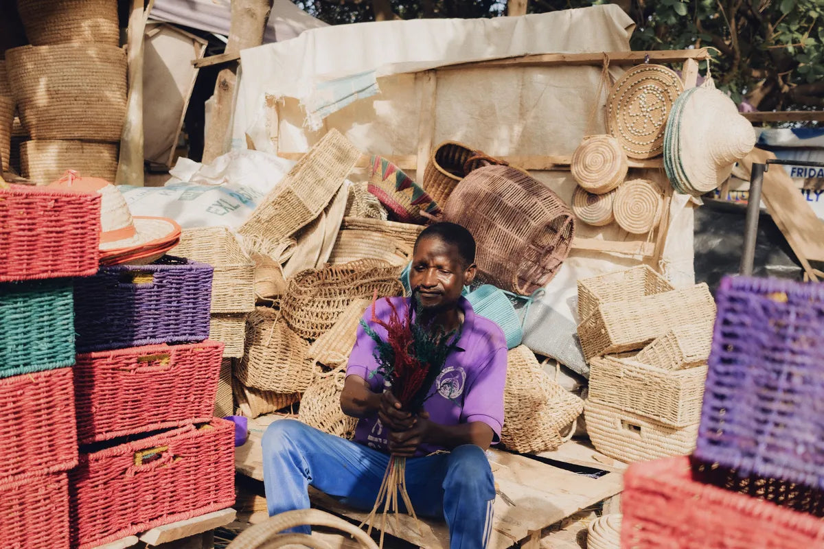 A basket seller seated among stacks of handwoven baskets at an open-air market, surrounded by straw trays, storage baskets and dyed hampers.