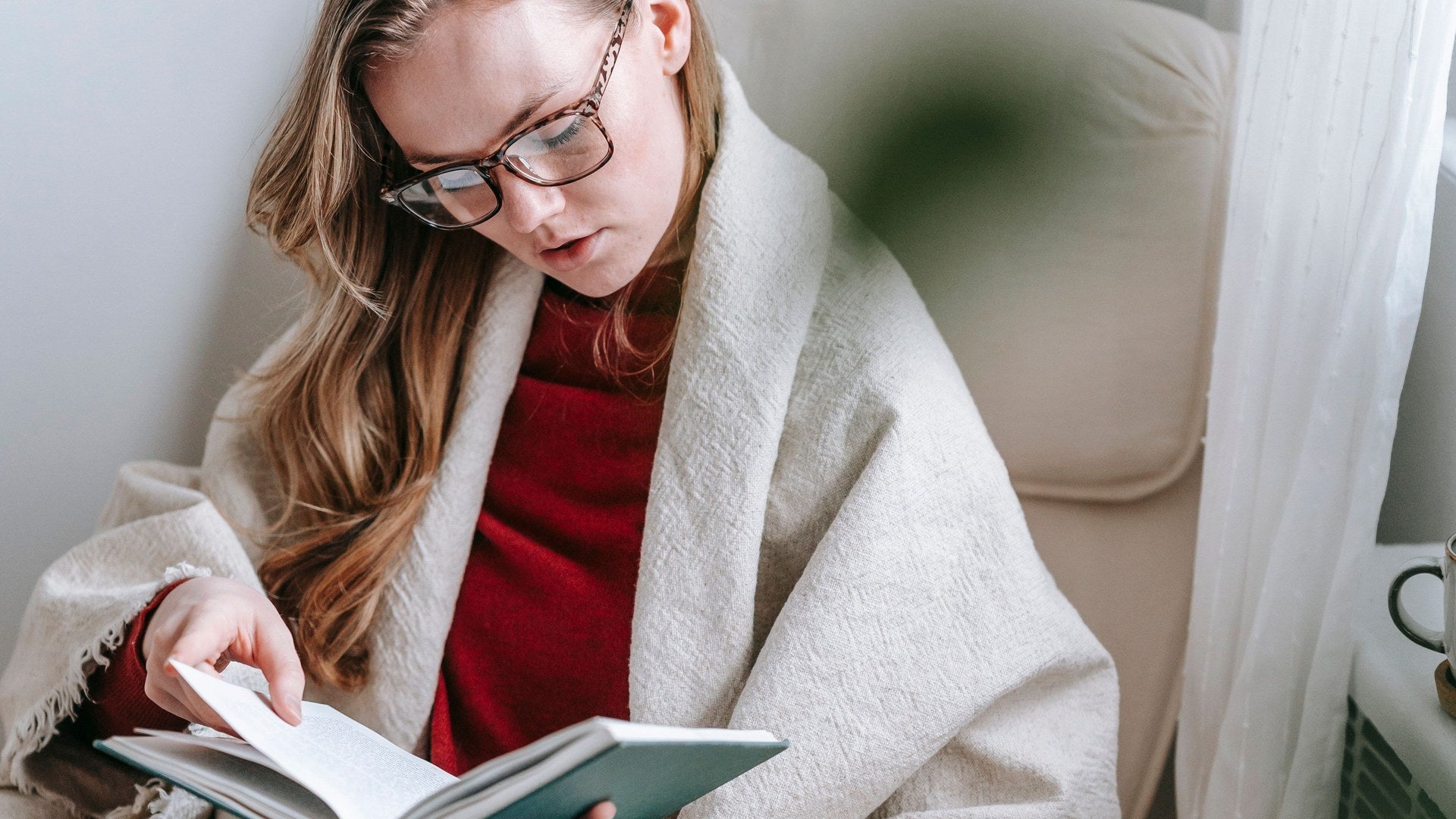 A woman in glasses reading in an armchair by a window, wrapped shoulders to lap in a cream handwoven throw with fringed edges.