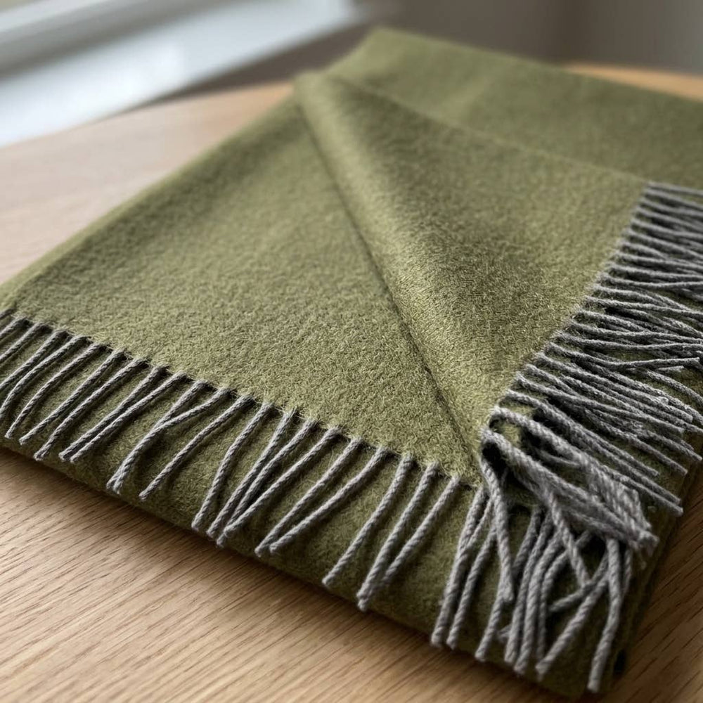 An olive baby alpaca throw folded on a light wood surface, showing the grey knotted fringe.