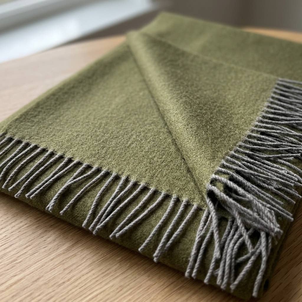 An olive baby alpaca throw folded on a light wood surface, showing the grey knotted fringe.