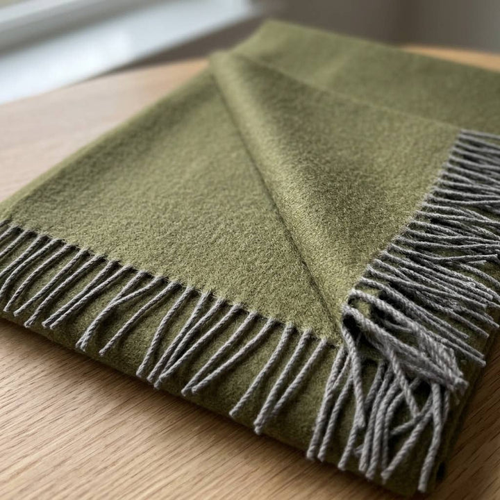 An olive baby alpaca throw folded on a light wood surface, showing the grey knotted fringe.