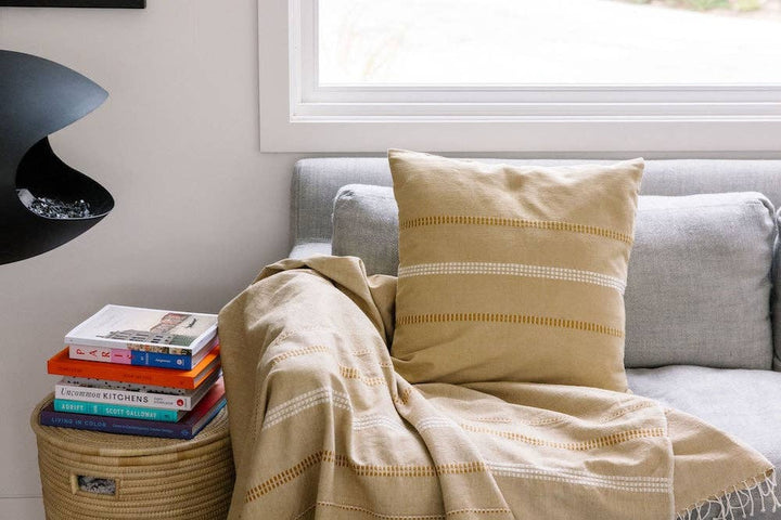 A handwoven cotton pillow cover and matching throw on a grey sofa beside a stack of books.