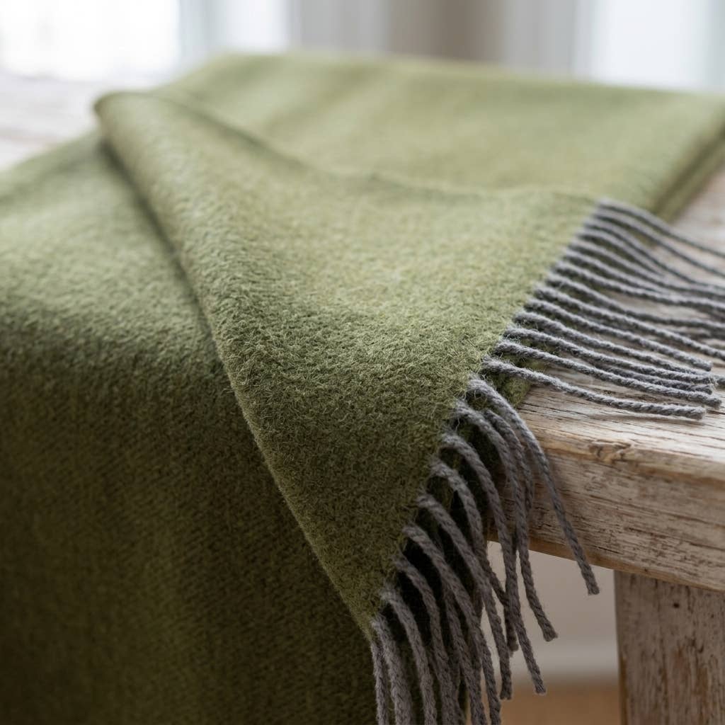 An olive baby alpaca throw draped over the edge of a weathered wooden table with grey fringe hanging.