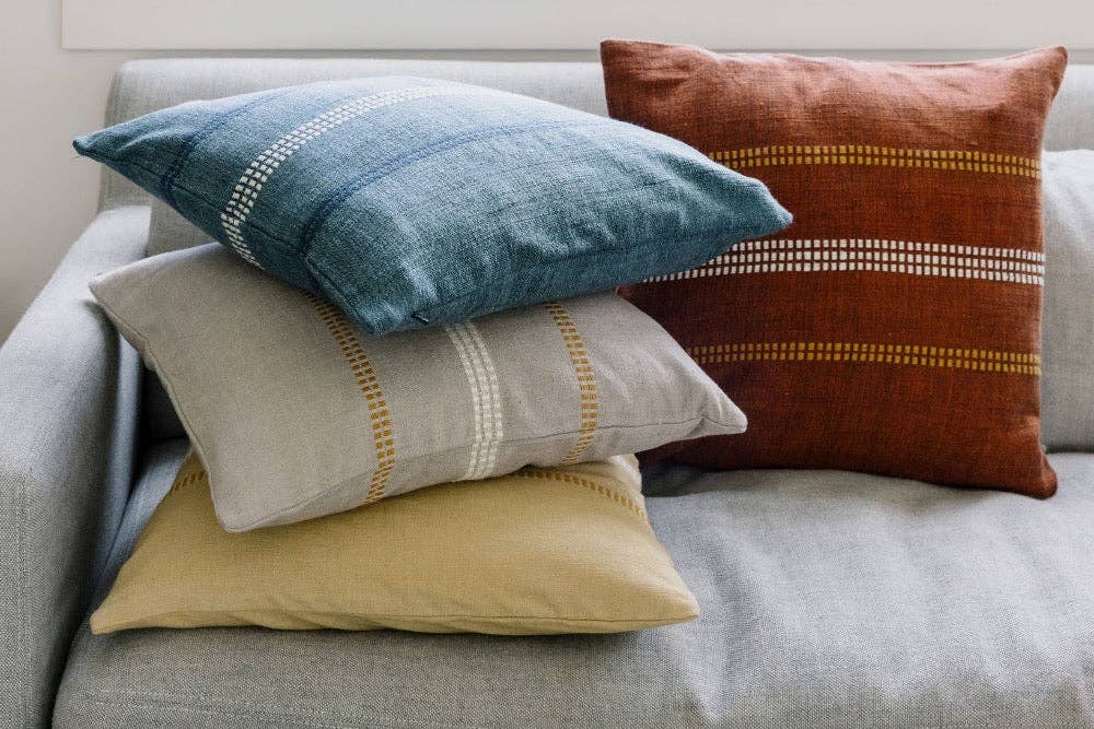 Four handwoven cotton pillow covers stacked on a grey sofa with the copper one on top.