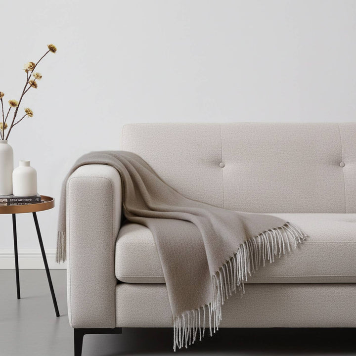 A hazelnut baby alpaca throw draped over the arm of a cream sofa with the fringe hanging at the front.
