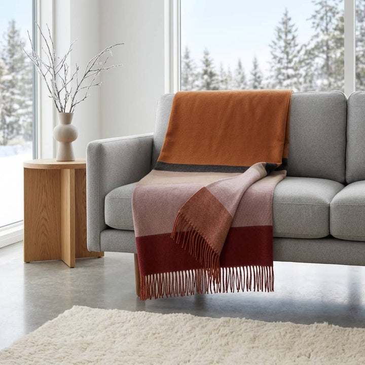 A fringed alpaca throw laid over the back of a grey sofa beside a window looking onto snow.