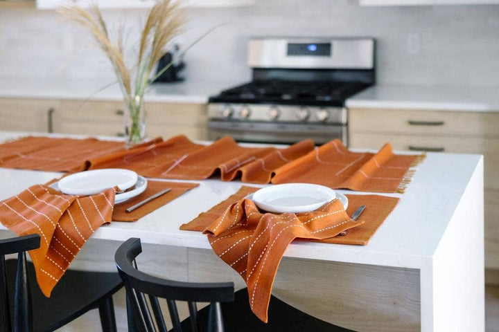 Woven cotton table linens in rust set along a white kitchen island with plates and napkins.