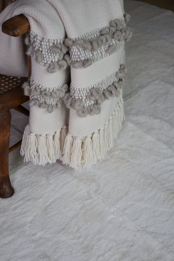 Ivory wool throw with grey bobbled bands folded over a wooden chair on a pale sheepskin rug.