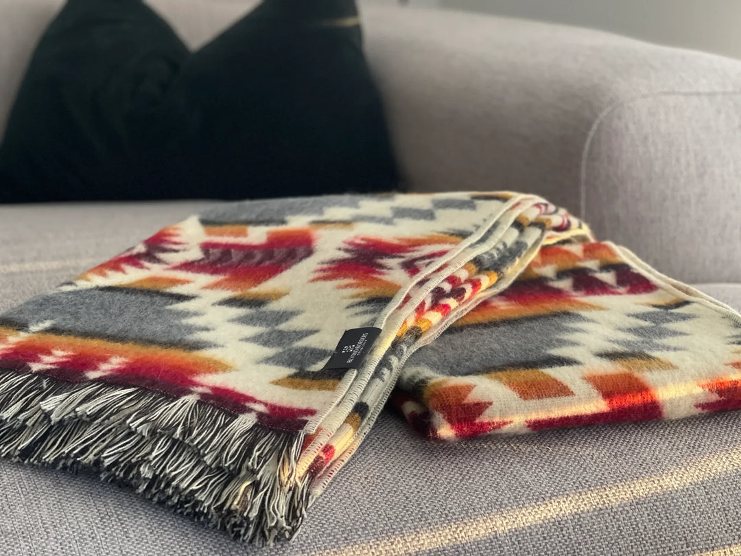 Campfire Ash fringe throw blanket folded on a grey sofa, showing crimson, orange, and charcoal pattern