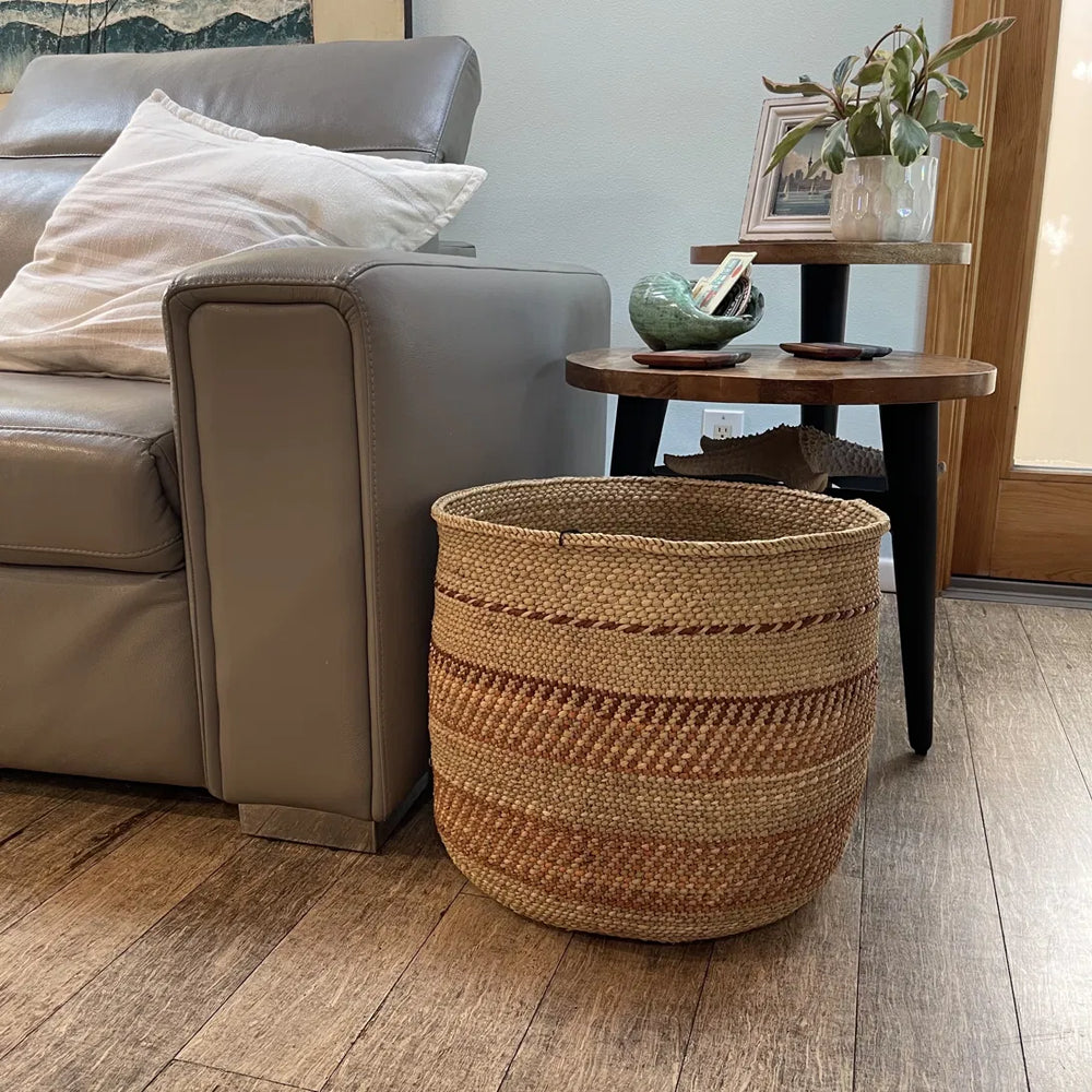 An Auburn Stripe extra large grass basket on a wood floor beside a leather sofa and round side table