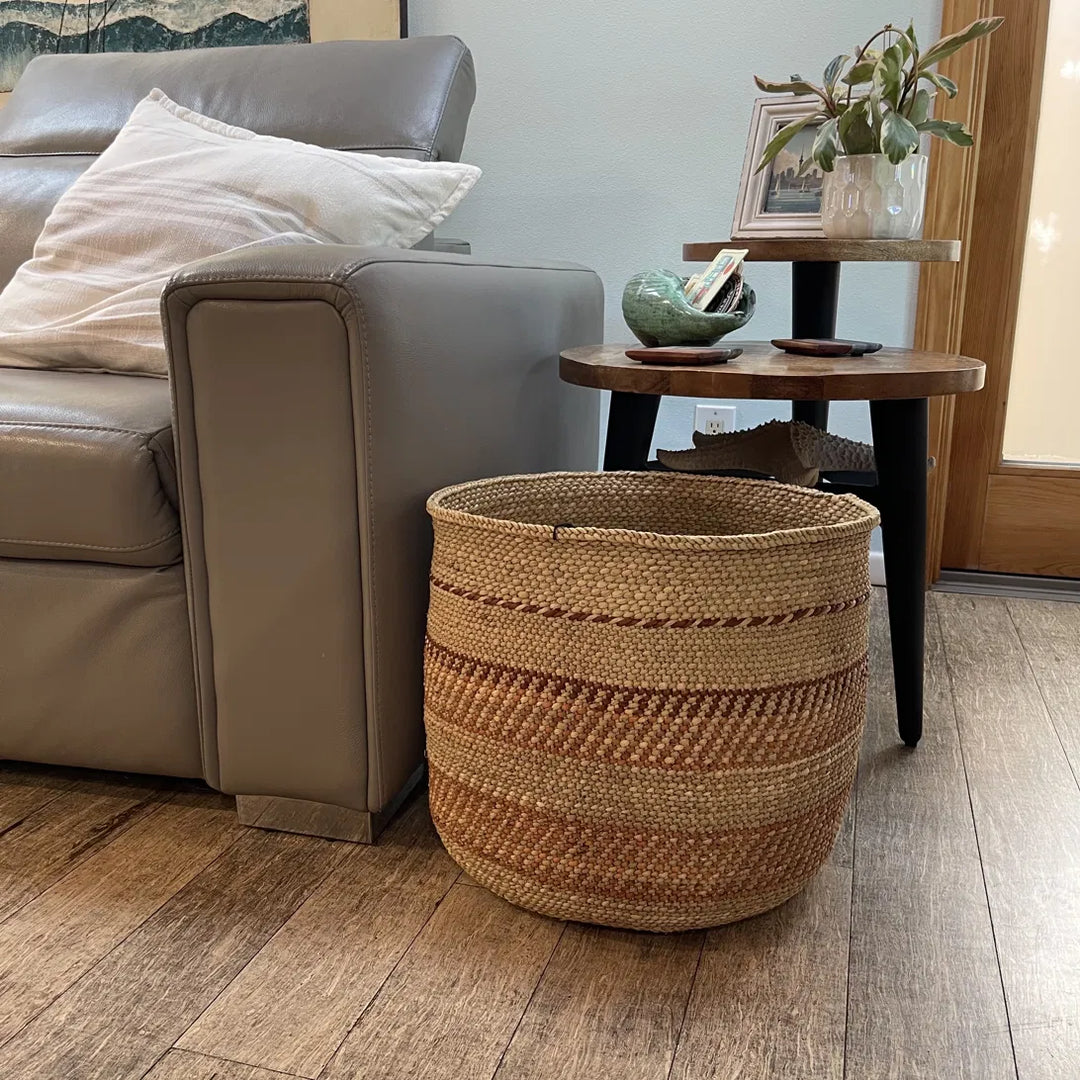 An Auburn Stripe extra large grass basket on a wood floor beside a leather sofa and round side table