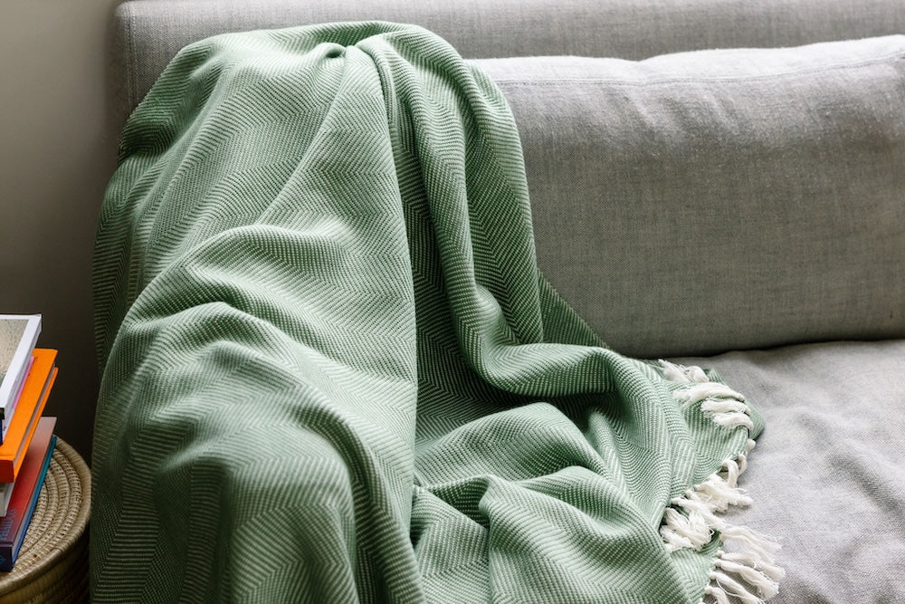 Mint herringbone cotton throw piled on a grey sofa beside a stack of books.