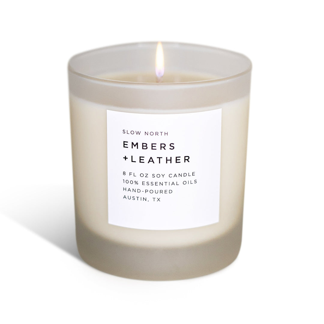 Embers &amp; Leather signature candle in a glass jar with textured leather label and warm flame.