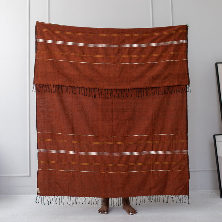 Copper cotton throw held up against a white wall, showing its full size, woven motif bands and twisted fringe.