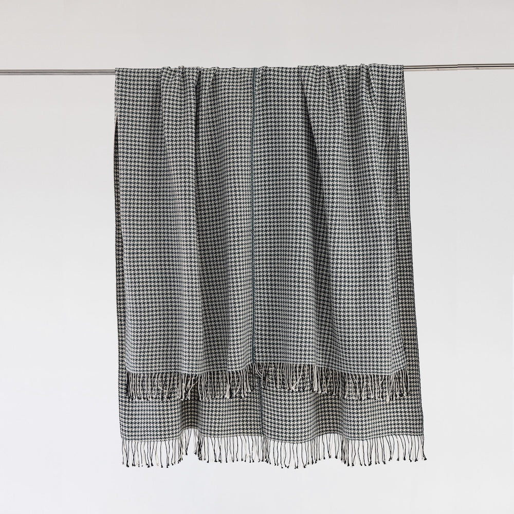 Slate and white pinwheel check cotton throw hanging from a metal rail against a pale wall, with hand-tied fringe.