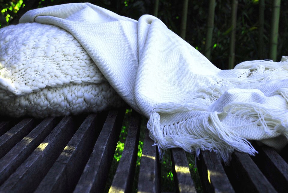 Ivory wool throw draped over a slatted wooden bench beside a knit cushion.