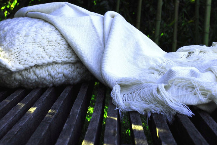 Ivory wool throw draped over a slatted wooden bench beside a knit cushion.
