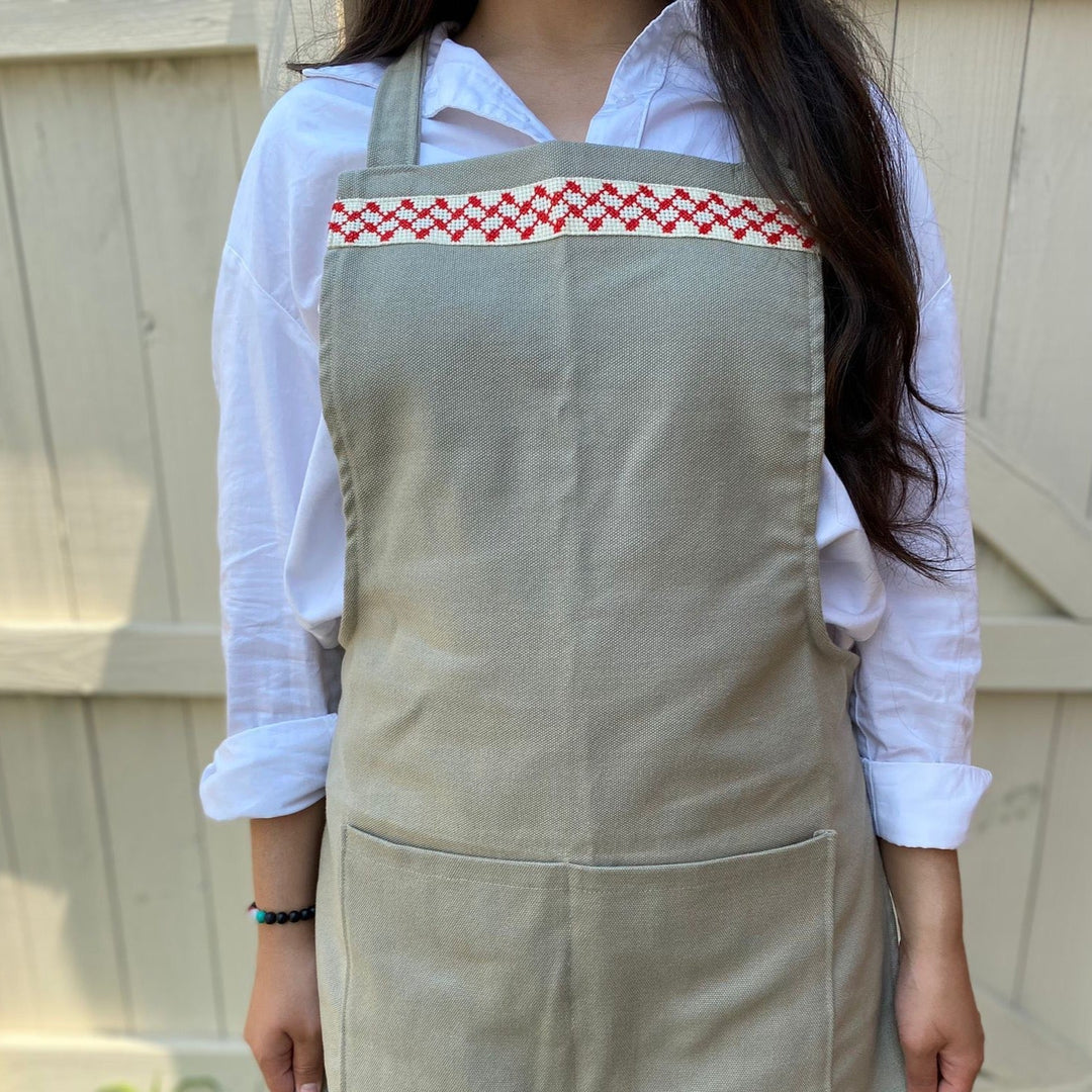 Close-up of the red-and-white kuffiyeh cross-stitch band on a sage-green apron.