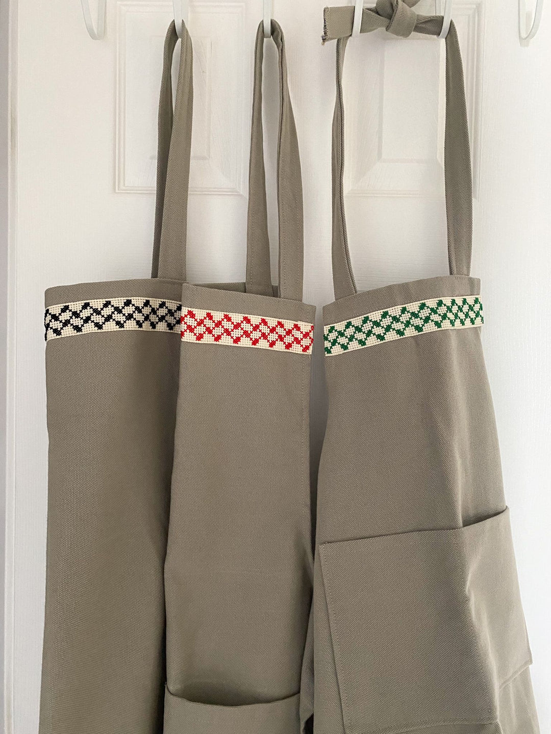 Full-length sage-green kitchen apron with a hand-embroidered red-and-white Palestinian kuffiyeh band.