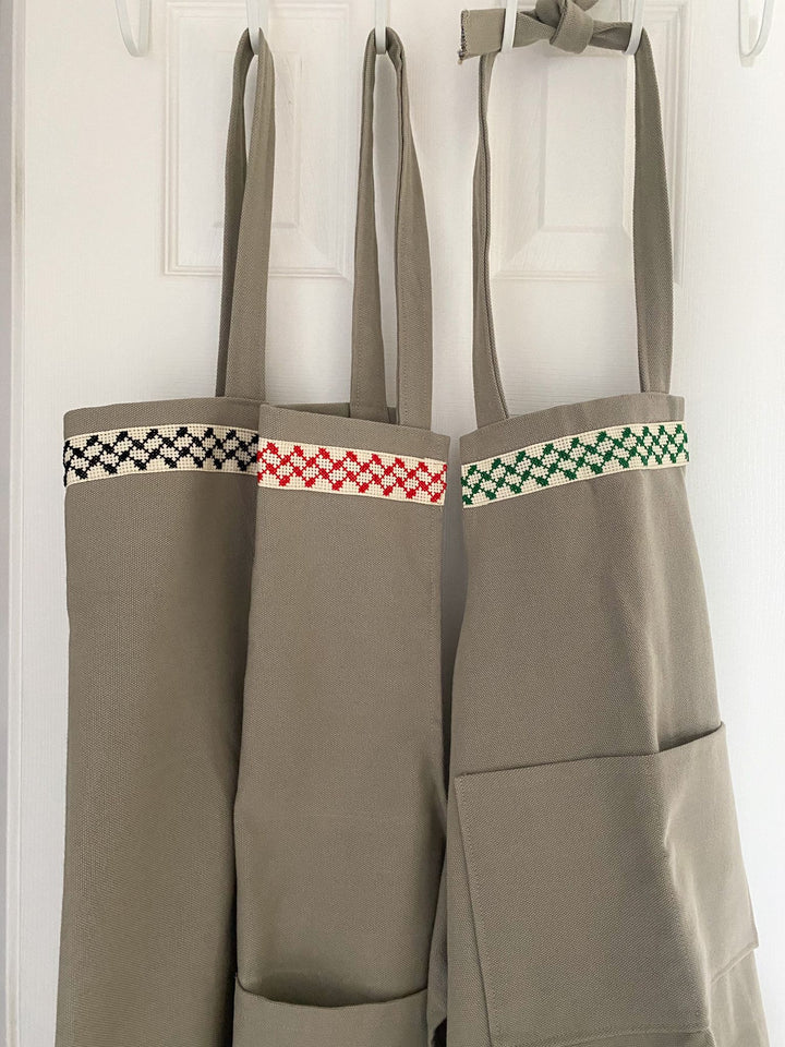 Full-length grey kitchen apron with a hand-embroidered black-and-white Palestinian kuffiyeh band.