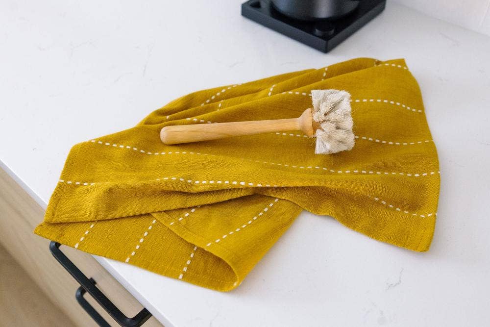 A mustard cotton tea towel folded on a white counter with a wooden dish brush.