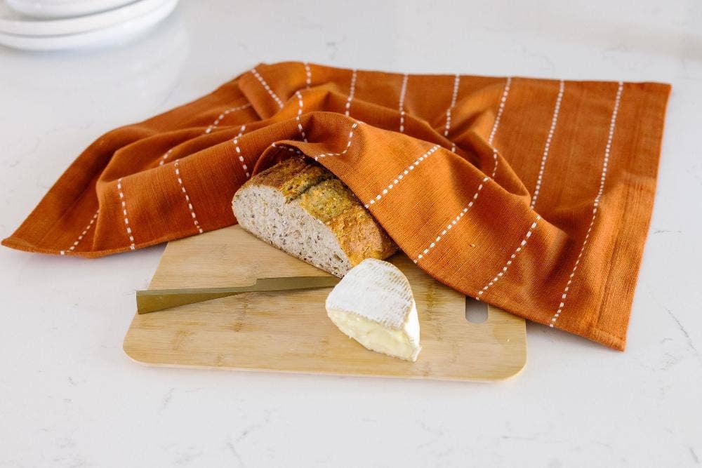 A rust cotton tea towel wrapped around a loaf of bread on a wooden board.