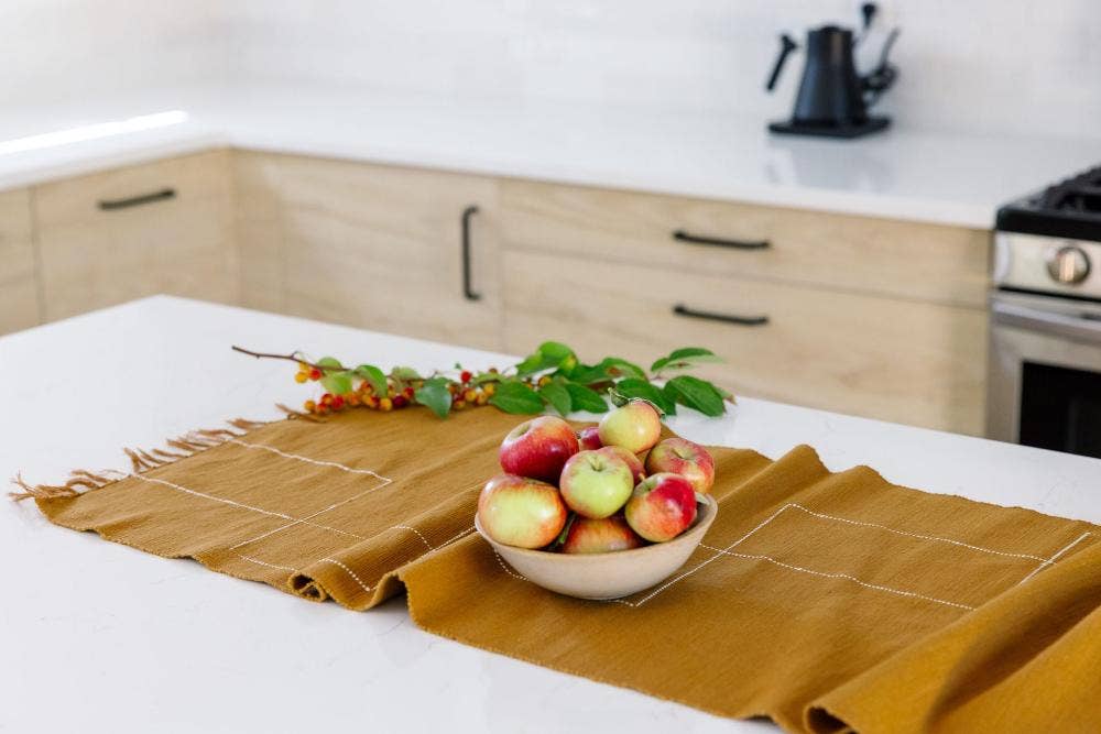 An ochre cotton table runner on a kitchen island with a bowl of apples and a berry branch.