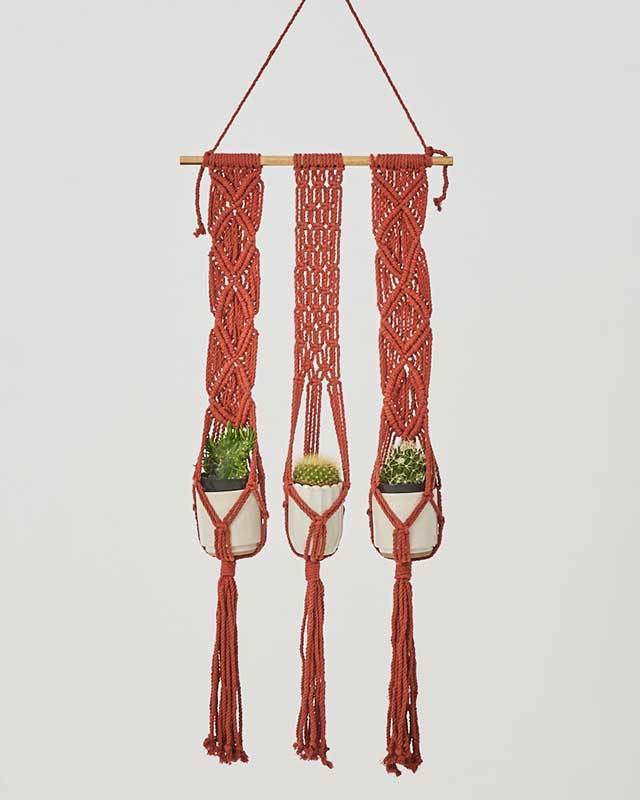 Tile-red three-tier macrame plant holder with intricate knotwork, hanging on a wall.
