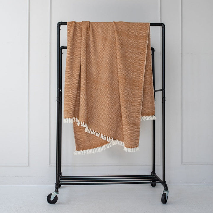 Bronze herringbone cotton throw hanging on a black clothes rail against a white wall, with natural white fringe.