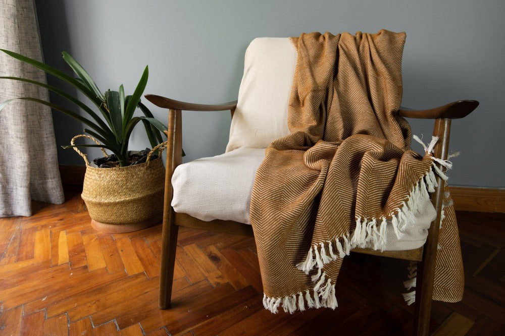 Bronze herringbone cotton throw draped over a wooden armchair on a parquet floor beside a potted plant.