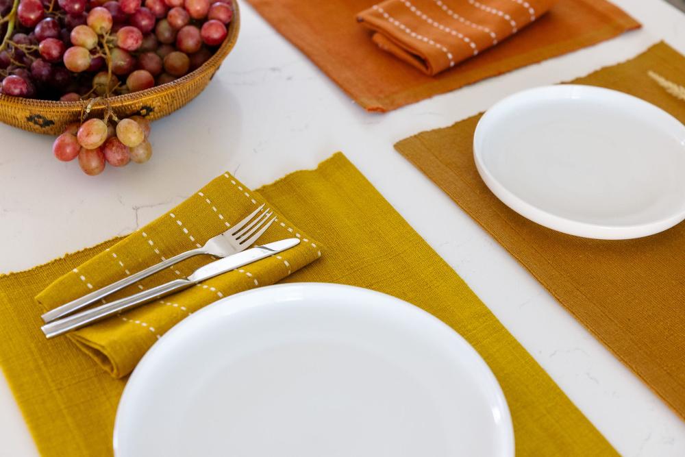 Gold cocktail napkins on handwoven placemats with white plates and a bowl of grapes