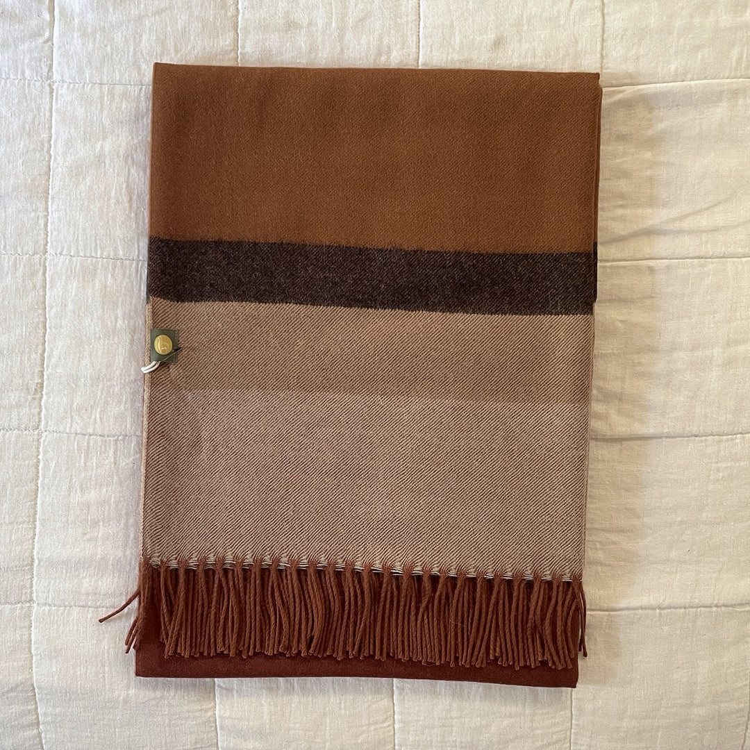 A folded woven scarf with horizontal stripes in brown, beige, and dark brown, finished with fringe along one edge and a small metal tag, laid flat on a white textured fabric background.