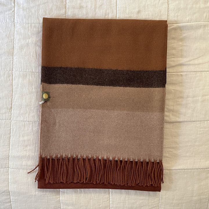A folded woven scarf with horizontal stripes in brown, beige, and dark brown, finished with fringe along one edge and a small metal tag, laid flat on a white textured fabric background.