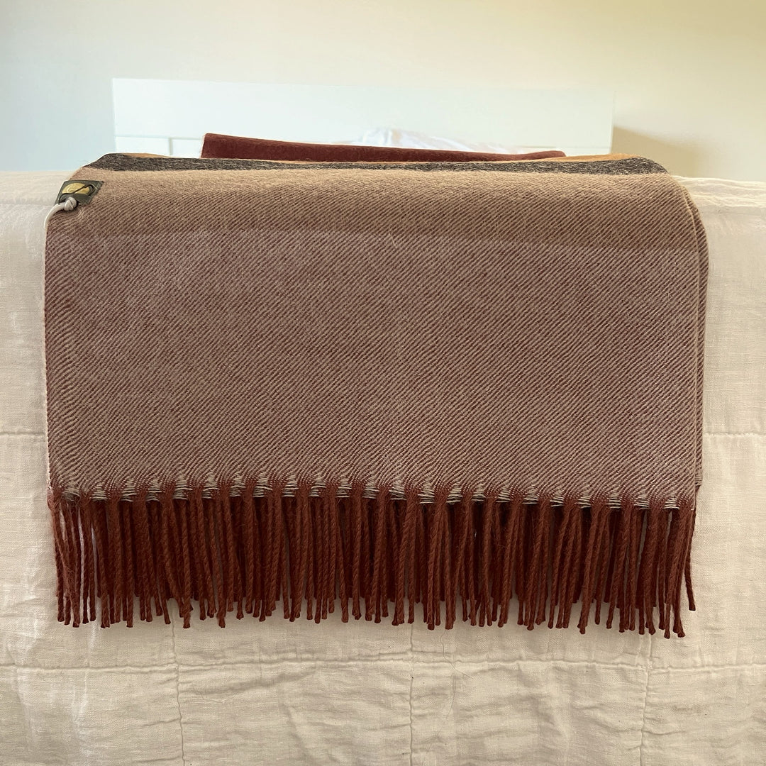 A brown and beige herringbone woven throw blanket with dark brown fringe, draped over a white quilted bed or sofa.