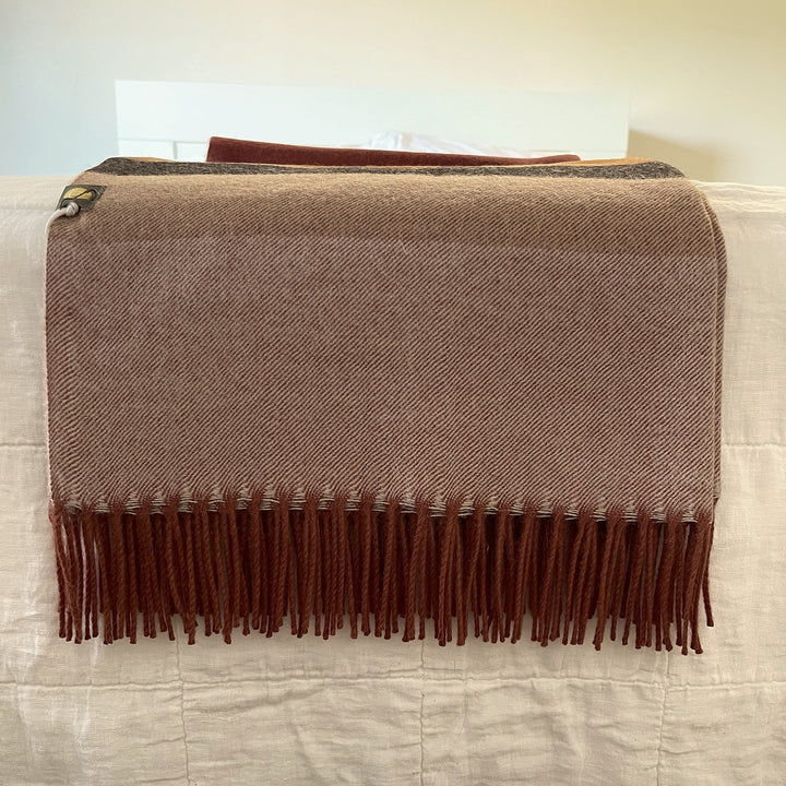 A brown and beige herringbone woven throw blanket with dark brown fringe, draped over a white quilted bed or sofa.