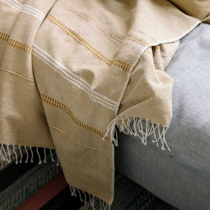 Almond cotton throw with gold woven bands draped over the arm of a grey sofa.