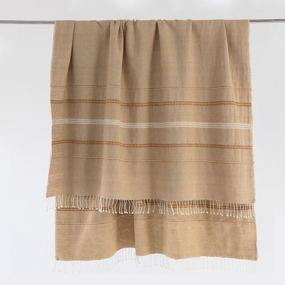 Almond cotton throw hanging from a rail, with gold and cream woven bands and twisted fringe.