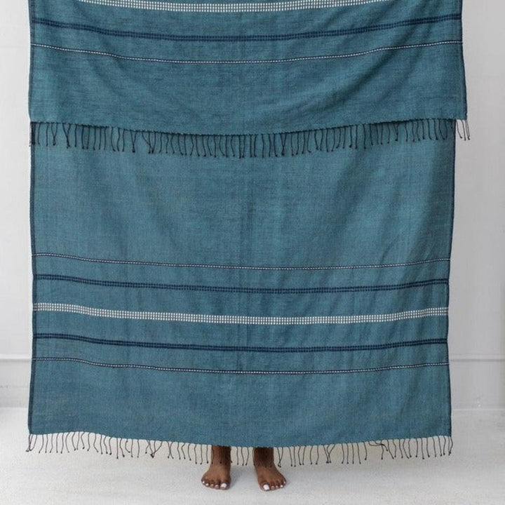 Steel blue cotton throw held up against a white wall, with navy and white woven bands.