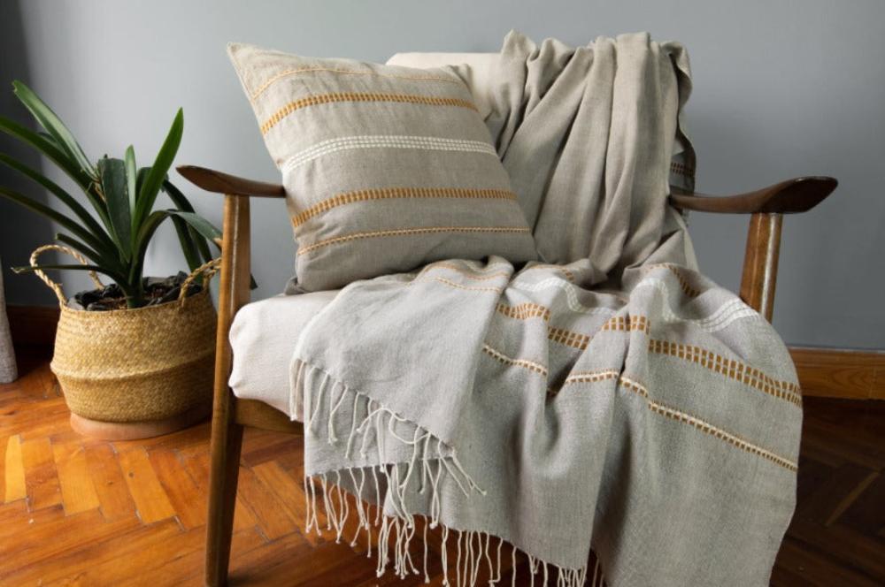 Stone cotton throw with copper accents draped over a wooden armchair beside a matching pillow and a potted plant.