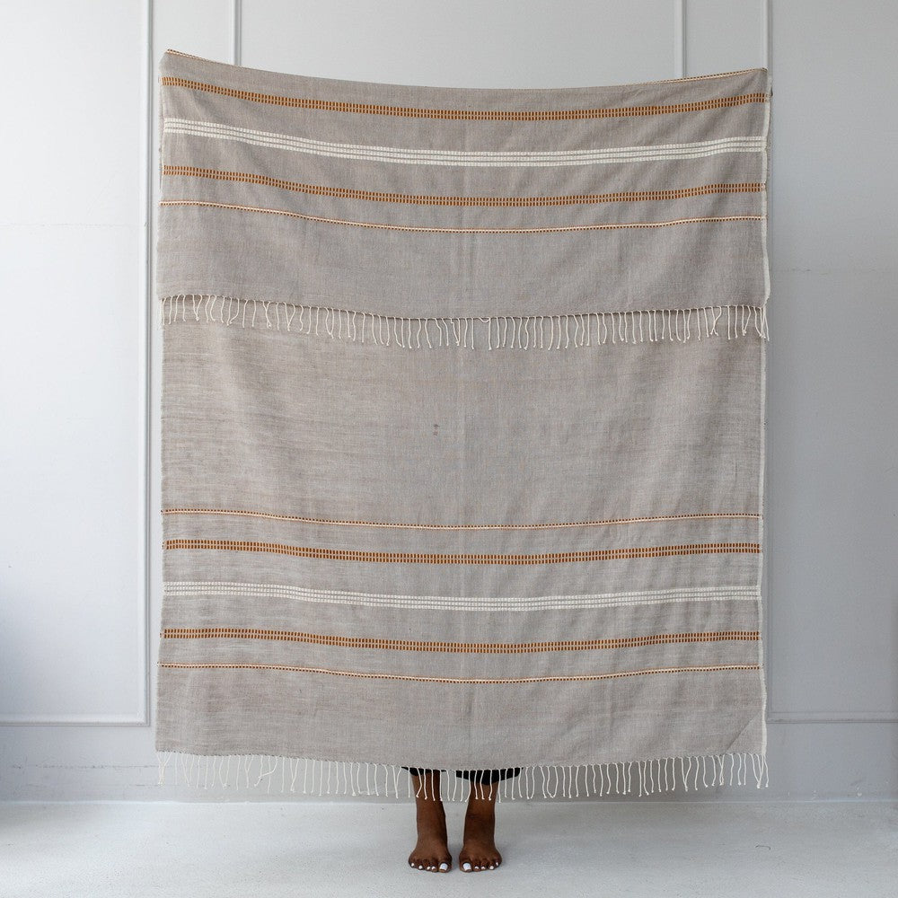 Stone cotton throw held up against a white wall, showing its copper and gold motif bands.