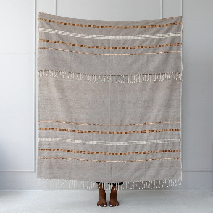 Stone cotton throw held up against a white wall, showing its copper and gold motif bands.